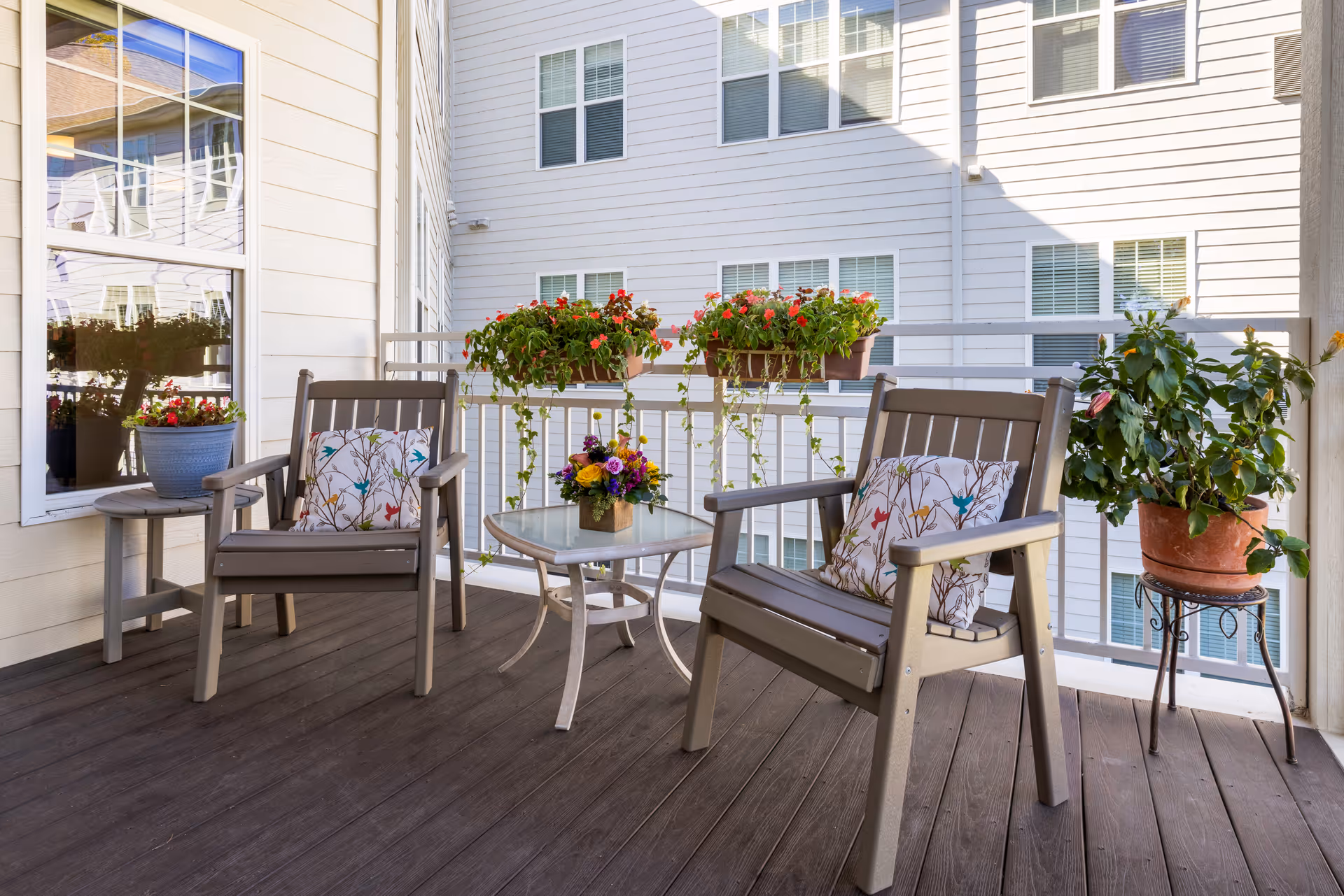 A cozy outdoor balcony area with two wooden chairs featuring floral patterned cushions, a small glass-top table with a flower arrangement, and several potted plants including hanging flower boxes on the railing. The balcony overlooks a white building with multiple windows.