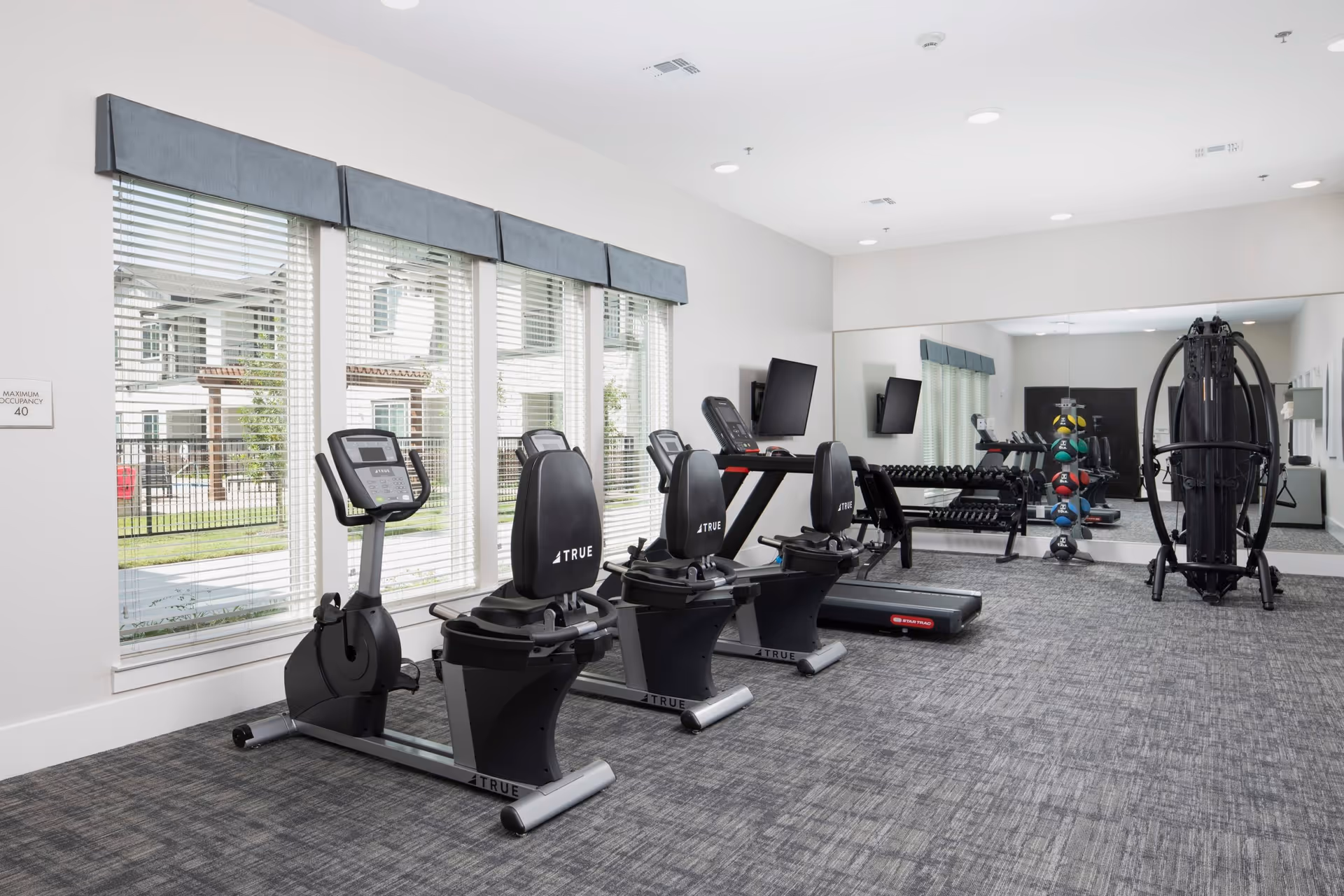A fitness room with exercise equipment including stationary bikes, a treadmill, a multi-function weight machine, a rack of dumbbells, and a rack of colorful medicine balls. Large windows with blinds allow natural light to enter, and a large mirror covers one wall.