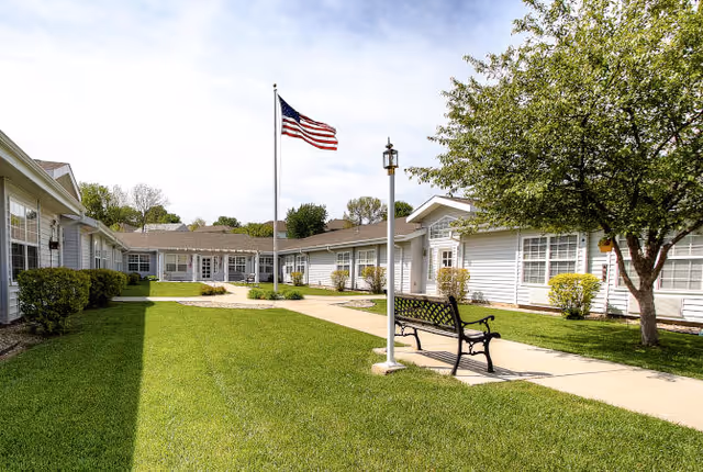 Outdoor courtyard area of a senior living facility with a well-maintained lawn, a paved walkway, a black metal bench, an American flag on a flagpole, and white single-story buildings surrounding the courtyard under a partly cloudy sky.