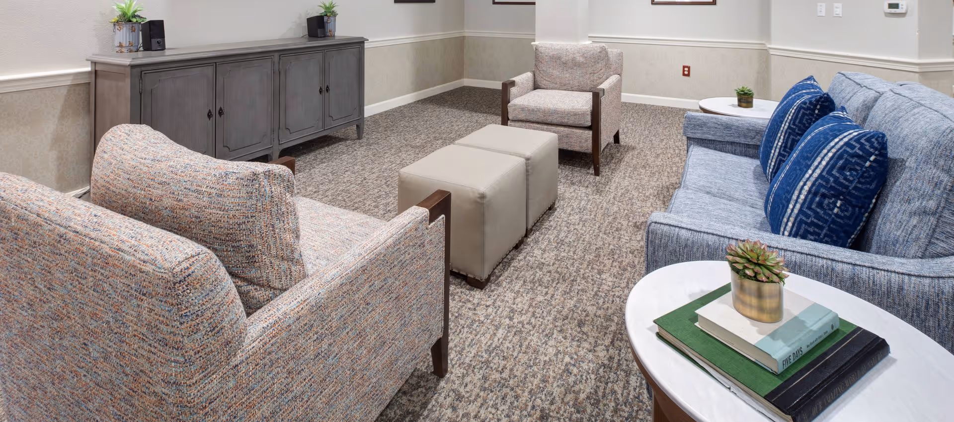 Carpeted lounge area with a blue sofa, patterned armchairs, two ottomans, side tables with books and a small plant, and a storage cabinet.