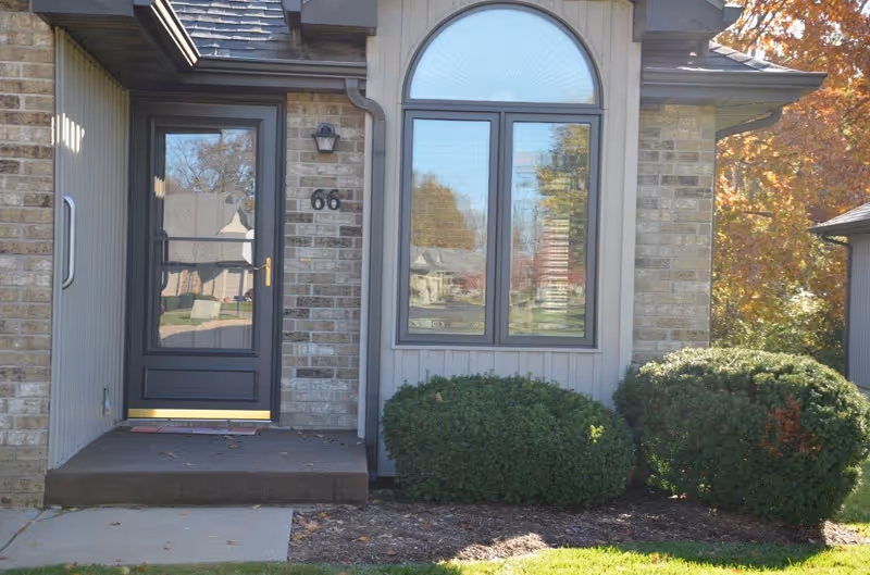 Front entrance of a brick-faced unit with a glass storm door numbered 66, an arched window, and shrubs along the foundation.