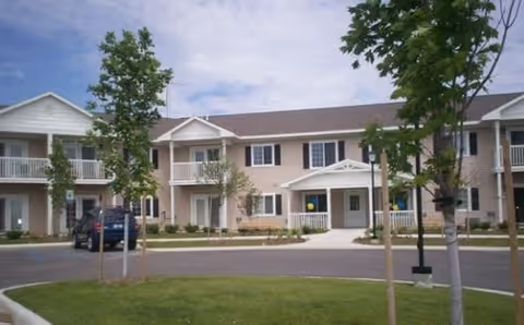 Exterior view of a two-story residential building with balconies and a driveway in front. There are young trees planted along the driveway and a parked vehicle visible on the left side. The sky is partly cloudy.