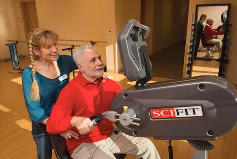 An elderly man in a red shirt is using a SciFit exercise machine in a fitness room, assisted by a smiling woman with blonde hair in a braid wearing a teal top. In the background, there is a mirror reflecting the man and woman, and exercise equipment is visible.