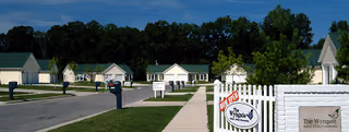A quiet residential street in a senior living community with single-story houses, mailboxes along the sidewalk, green lawns, and trees in the background under a clear blue sky. A white picket fence with a sign reading 'The Wyngate' is visible in the foreground.