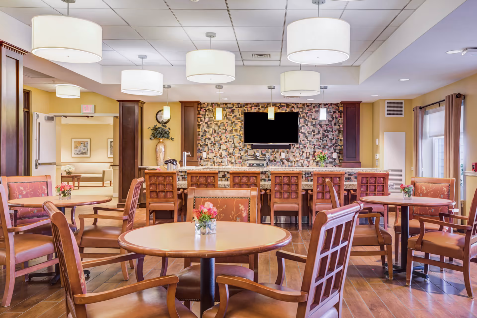 A bright and inviting dining area in a senior living facility featuring round wooden tables with floral centerpieces and cushioned chairs. In the background, there is a bar counter with high chairs, a mosaic tile wall, and a mounted flat-screen TV. The room is well-lit with multiple ceiling lights and has warm yellow walls and wooden flooring.