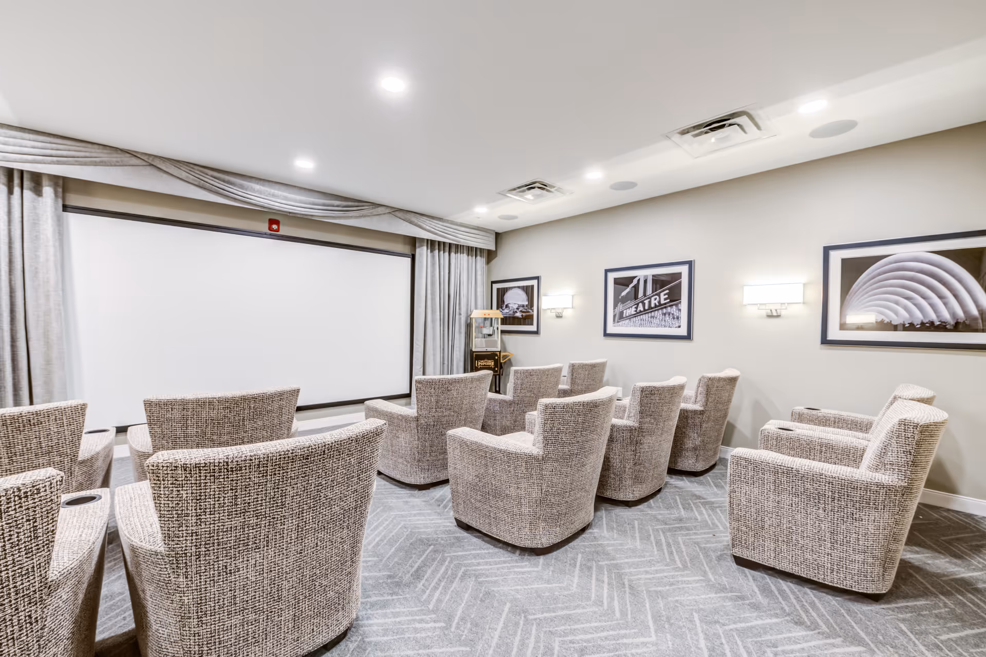 A small theater room with a large white projection screen at the front, surrounded by beige curtains. The room has two rows of comfortable upholstered armchairs arranged facing the screen. The walls are decorated with framed black and white photographs, and the room is well-lit with ceiling lights and wall sconces.