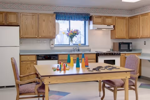 A kitchen area with wooden cabinets, a white refrigerator, stove, microwave, and dishwasher. A window above the sink has a blue valance and a vase of flowers on the windowsill. In the center, there is a wooden table with colorful cone-shaped objects and two chairs with purple upholstery.