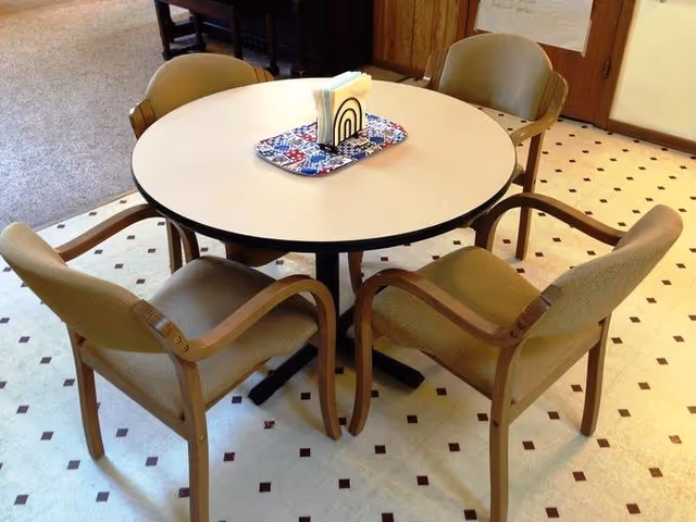 A round table with a light-colored top surrounded by four beige cushioned chairs with wooden armrests. On the table is a small tray holding napkins and a napkin holder. The floor has a white and brown diamond pattern, and part of a carpeted area and wooden door are visible in the background.