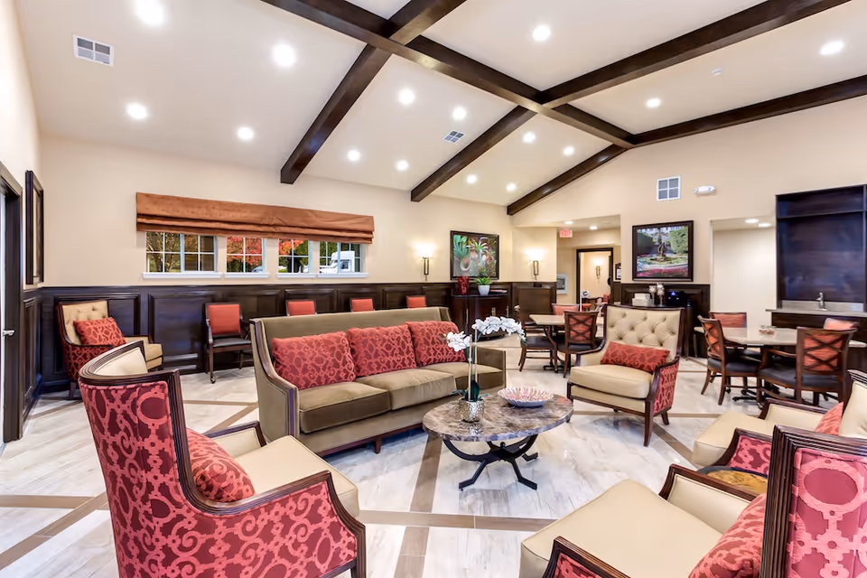 Bright communal lounge with sofas, patterned armchairs, a central coffee table and dining tables under exposed ceiling beams.