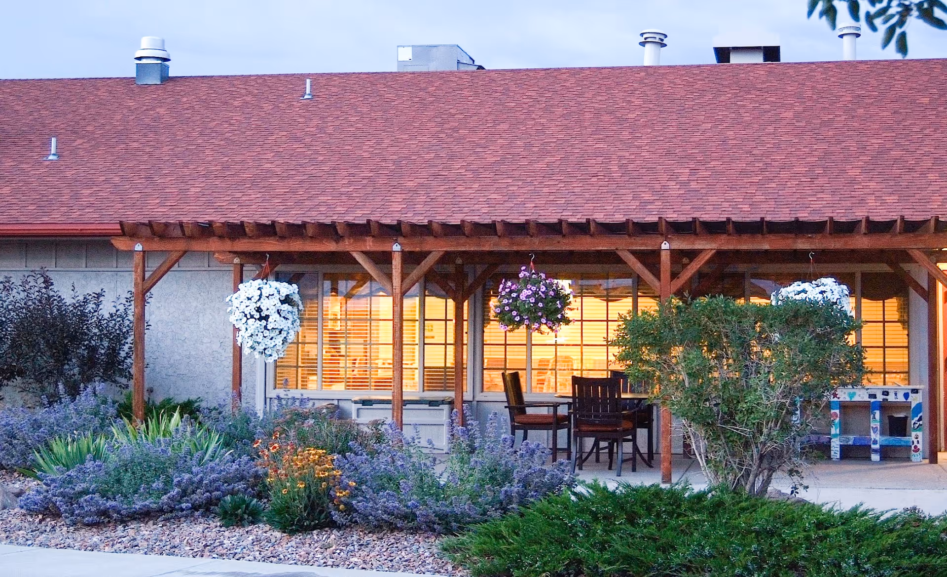 Outdoor patio area of San Luis Care Center with wooden pergola, hanging flower baskets, chairs and table, surrounded by various plants and shrubs in a landscaped garden.