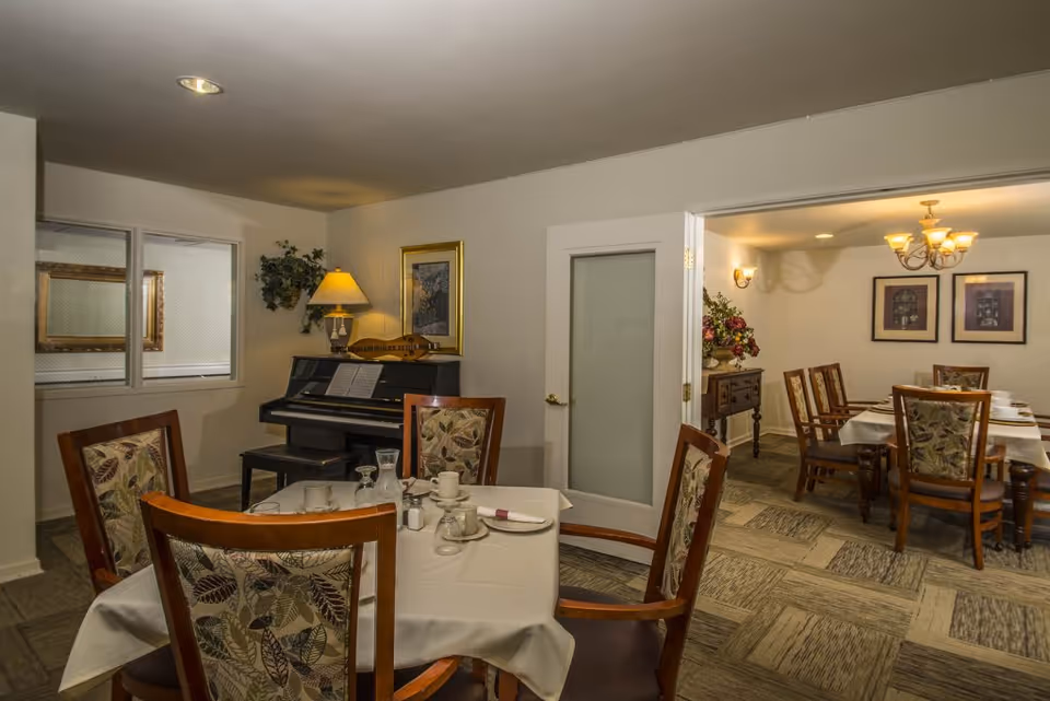Interior view of a dining area in an assisted living facility with tables covered in white tablecloths, set with cups, plates, and utensils. There are wooden chairs with patterned upholstery around the tables. A piano with sheet music and a lamp is against one wall, next to a window and a framed picture. Another dining area with a chandelier and framed artwork is visible through an open doorway.