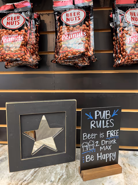 A display with hanging bags of Beer Nuts Original Peanuts above a black framed star decoration and a small chalkboard sign that reads 'PUB RULES Beer is FREE 2 Drink MAX Be Happy' on a marble countertop.