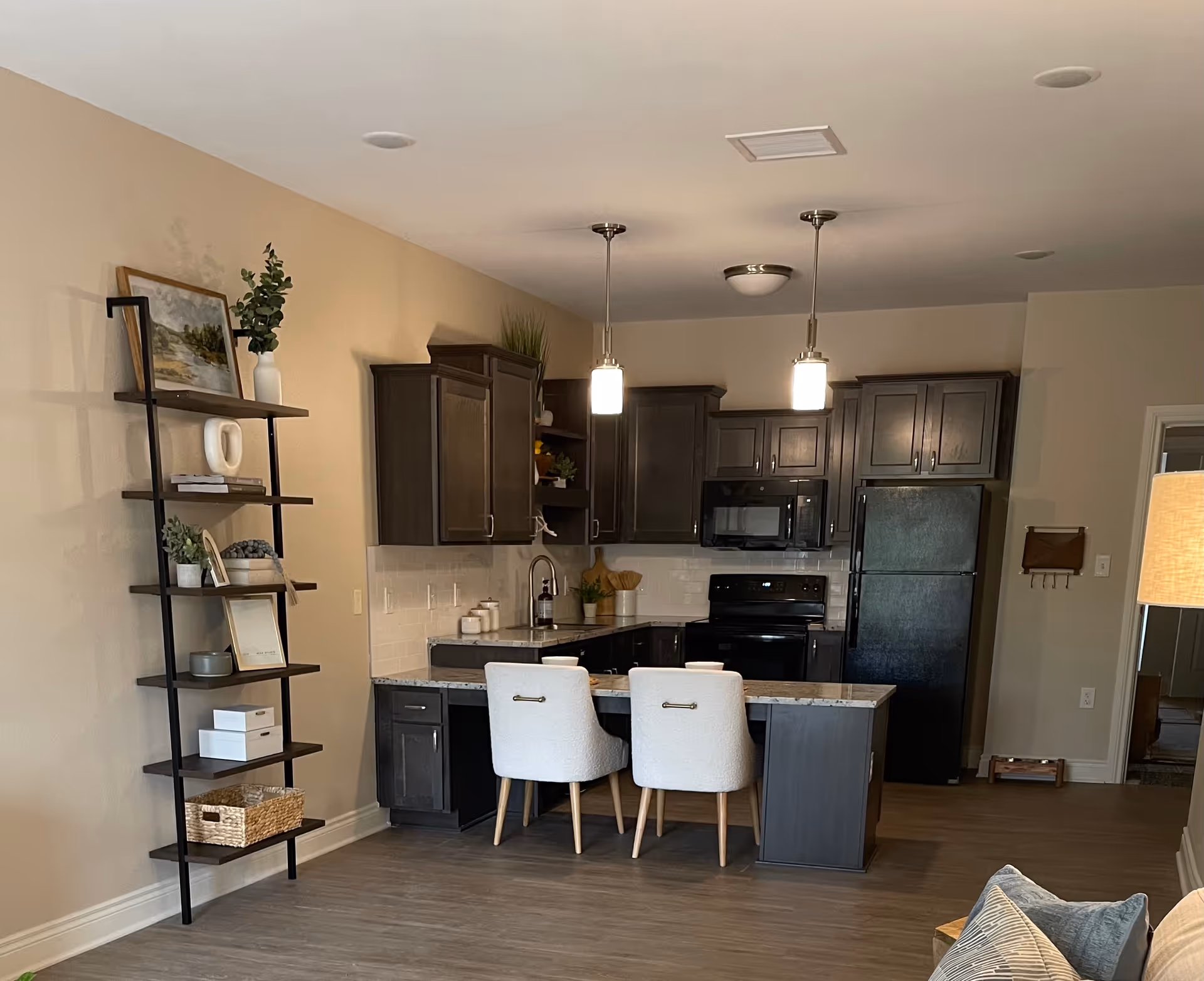 Modern kitchen area with dark wood cabinets, black appliances including a refrigerator, stove, and microwave. A kitchen island with a granite countertop has two white upholstered chairs. To the left, there is a tall black metal and wood shelving unit with decorative items and plants. The floor is wood, and the walls are painted beige.