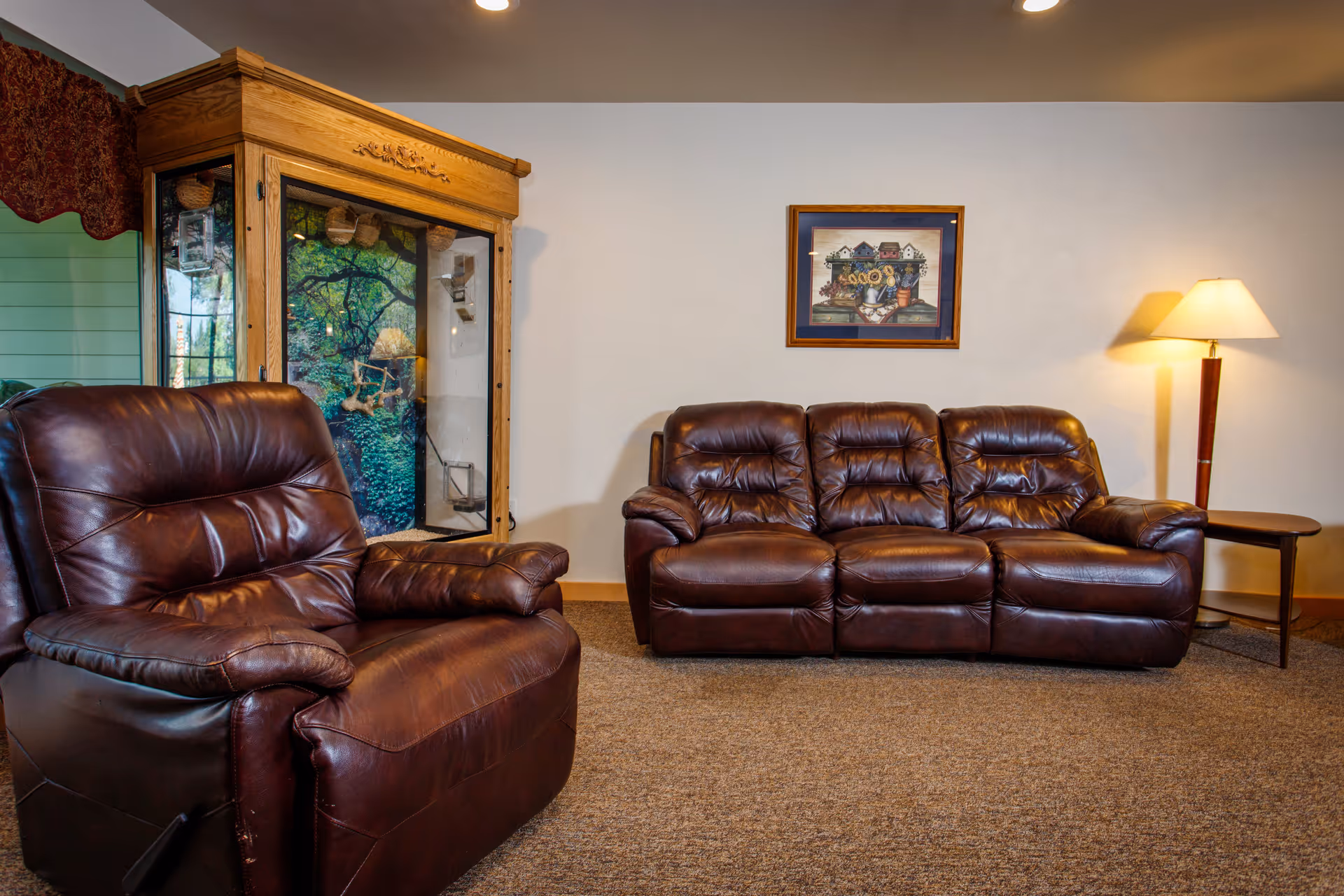 A cozy sitting area with a brown leather recliner and three-seat leather sofa, a side table with lamp, framed artwork on the wall, and a wooden display cabinet.