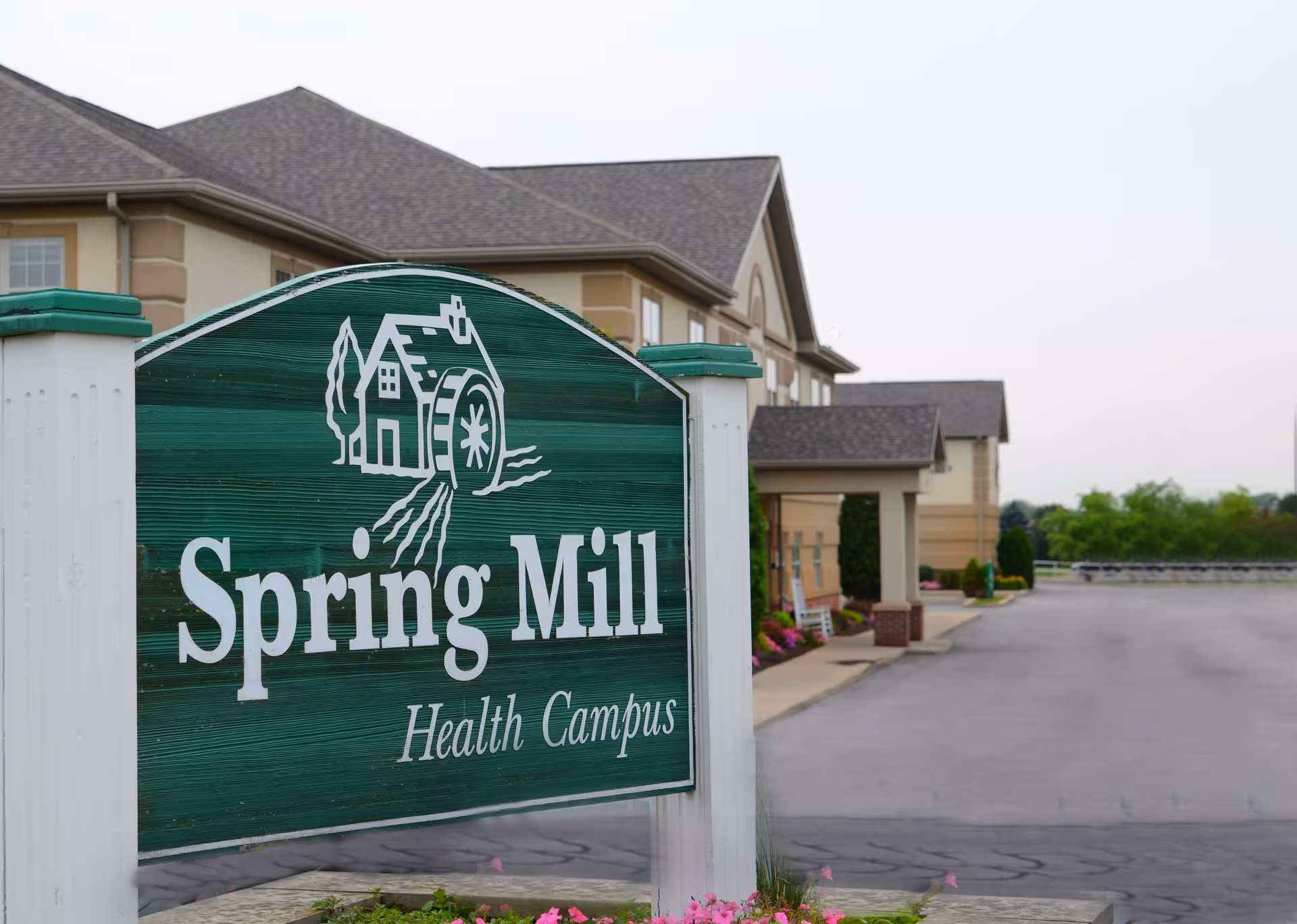 A green wooden sign with white lettering that reads 'Spring Mill Health Campus' in front of a beige building with a gray roof and a paved driveway, surrounded by some greenery and flowers.
