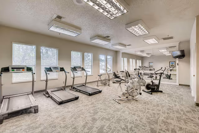 A well-lit fitness room with several treadmills, stationary bikes, and exercise equipment arranged neatly. The room has large windows with blinds, carpeted flooring, and fluorescent ceiling lights. A wall-mounted TV is visible in the corner.