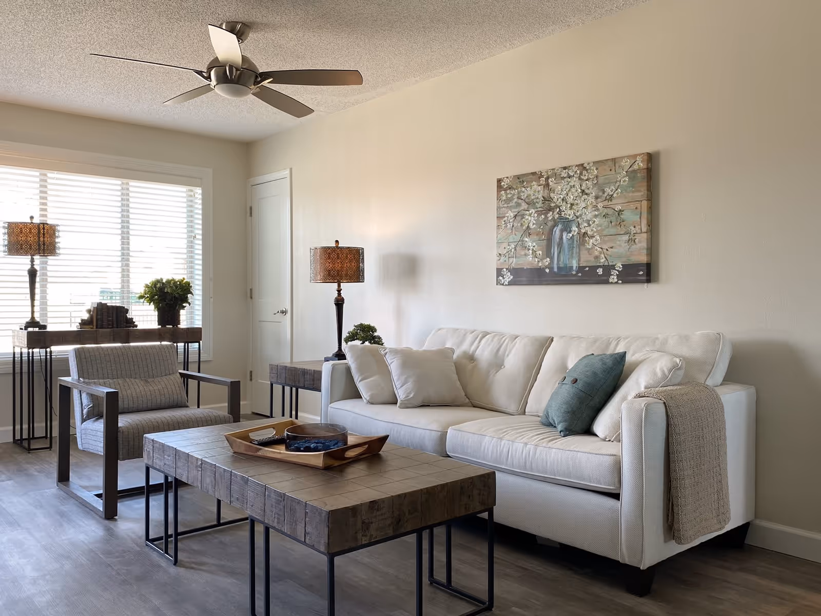 A bright and cozy living room with a white sofa adorned with several cushions and a beige throw blanket. In front of the sofa is a rustic wooden coffee table with a tray holding decorative items. To the left, there is a modern armchair with a cushion, a side table with a lamp, and a console table with books, a plant, and another lamp. A ceiling fan is mounted on the ceiling, and a large window with blinds allows natural light to fill the room. A floral painting hangs on the wall above the sofa.