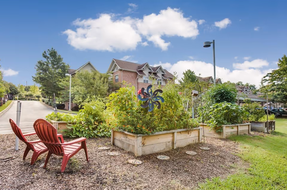 Outdoor garden area with raised wooden planter boxes filled with green plants and flowers. Two red Adirondack chairs are placed on a gravel surface facing the garden. In the background, there are trees, a street, and a multi-story brick building under a partly cloudy blue sky.