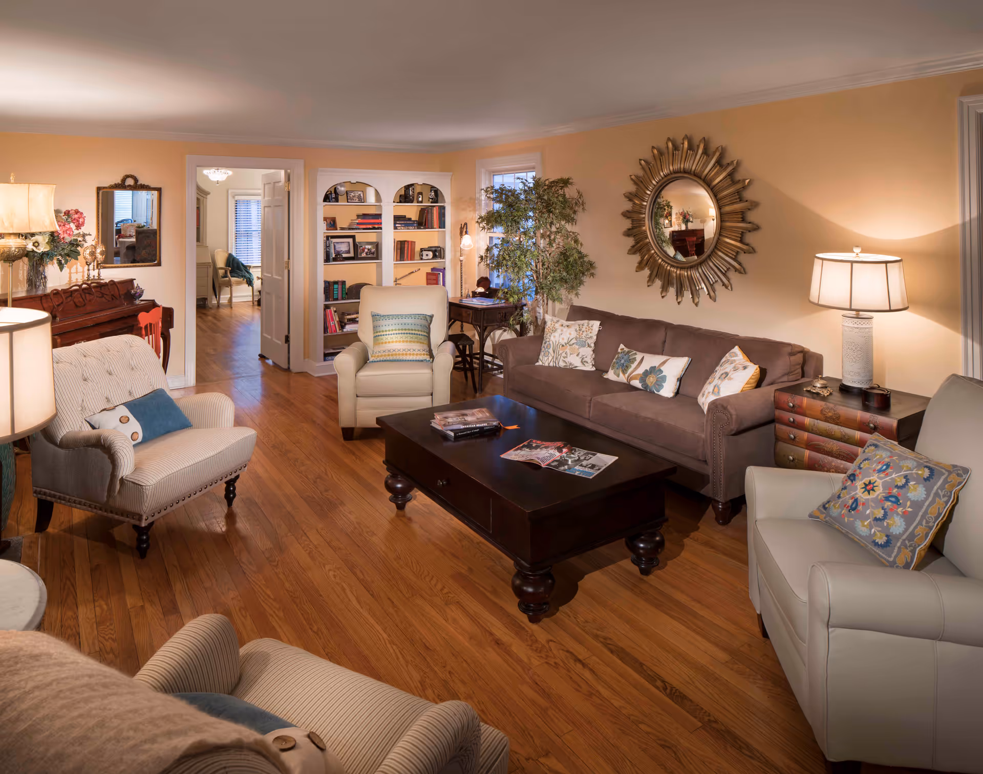 Warm, well-decorated living room with sofas and armchairs around a central coffee table, bookshelf, and decorative mirror.