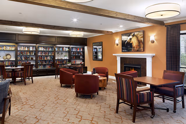 A cozy living room area with a fireplace, surrounded by red upholstered chairs and wooden tables. The room features a carpeted floor with a patterned design, wooden ceiling beams, and built-in bookshelves filled with books and decorative items. A painting of a horse hangs above the fireplace, and large windows with curtains allow natural light to enter the space.