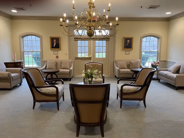A well-lit living room with beige walls and carpet, featuring a central chandelier with multiple lit candles. The room has two large windows and a glass door at the back, flanked by framed portraits on the walls. There are four beige upholstered armchairs arranged around a small table with a floral centerpiece, and three beige sofas positioned along the walls with side tables.