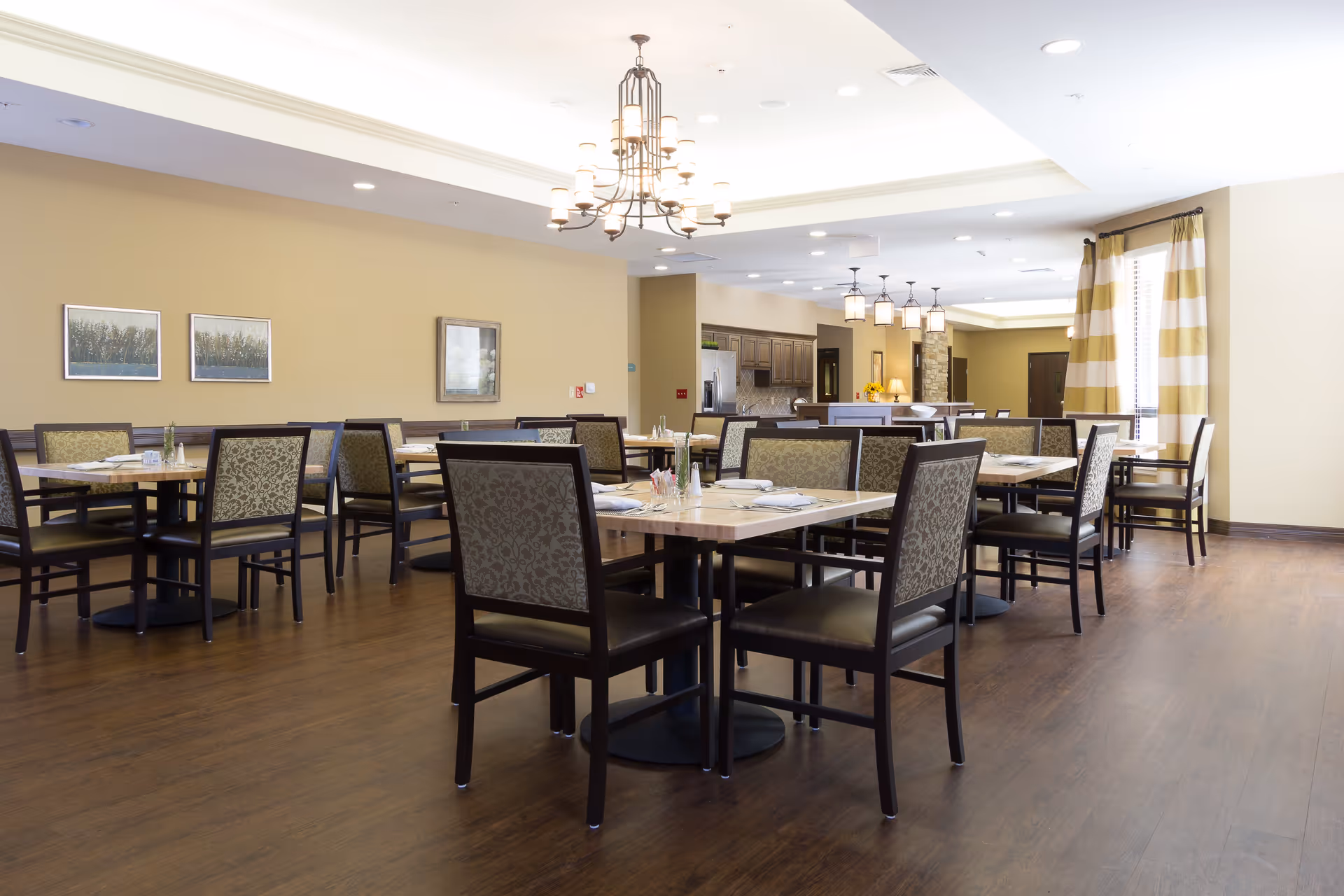 A spacious dining room with multiple tables and chairs arranged neatly. The room features wooden flooring, beige walls with framed artwork, large windows with striped curtains, and modern light fixtures hanging from the ceiling. In the background, there is a kitchen area with cabinets and appliances.