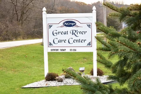 A white sign with decorative corners and the name 'McGregor Great River Care Center' displayed on a grassy area near a road, with trees and bushes in the background.