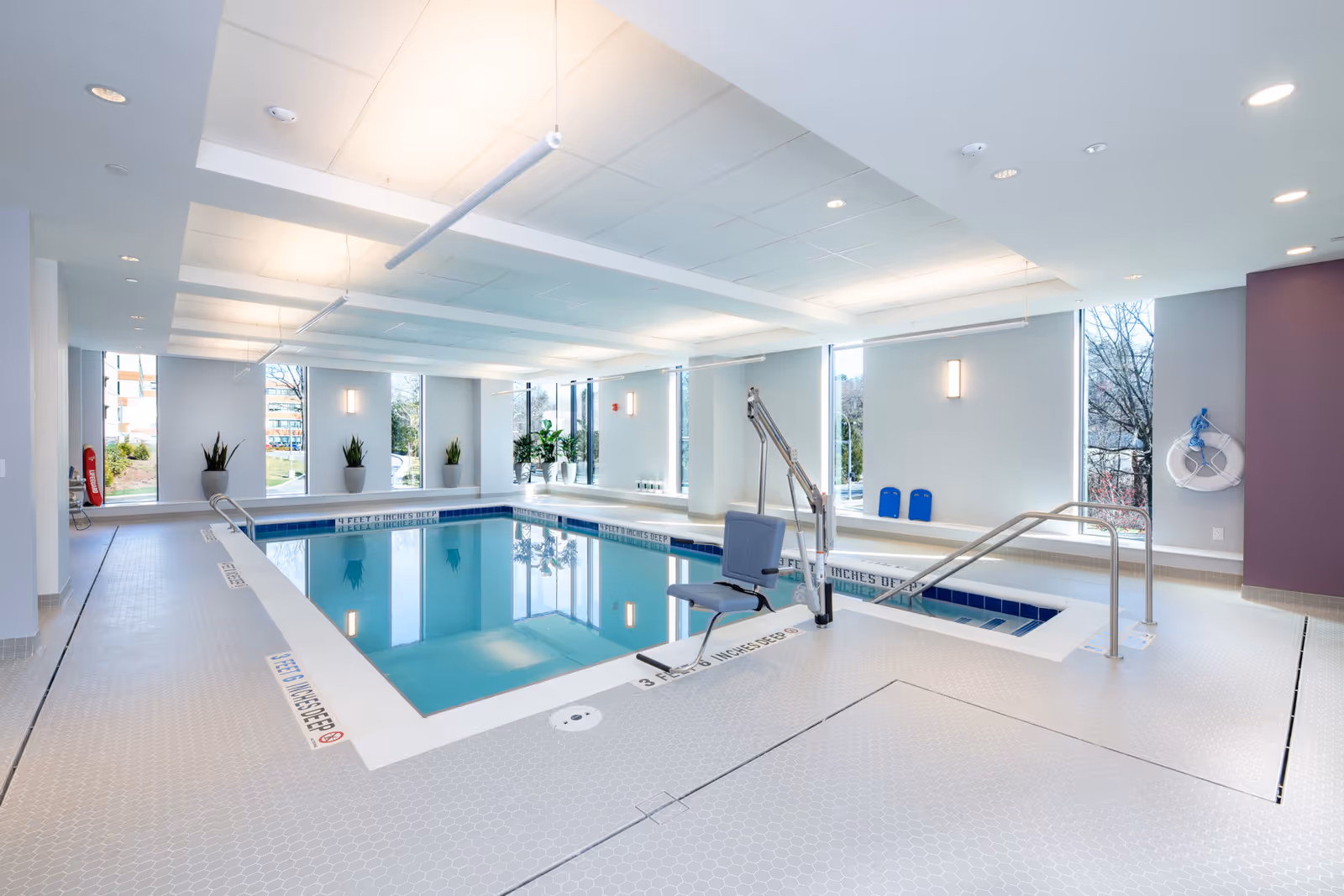 Bright indoor swimming pool with an accessibility lift, handrails, and large windows in a recreation room.