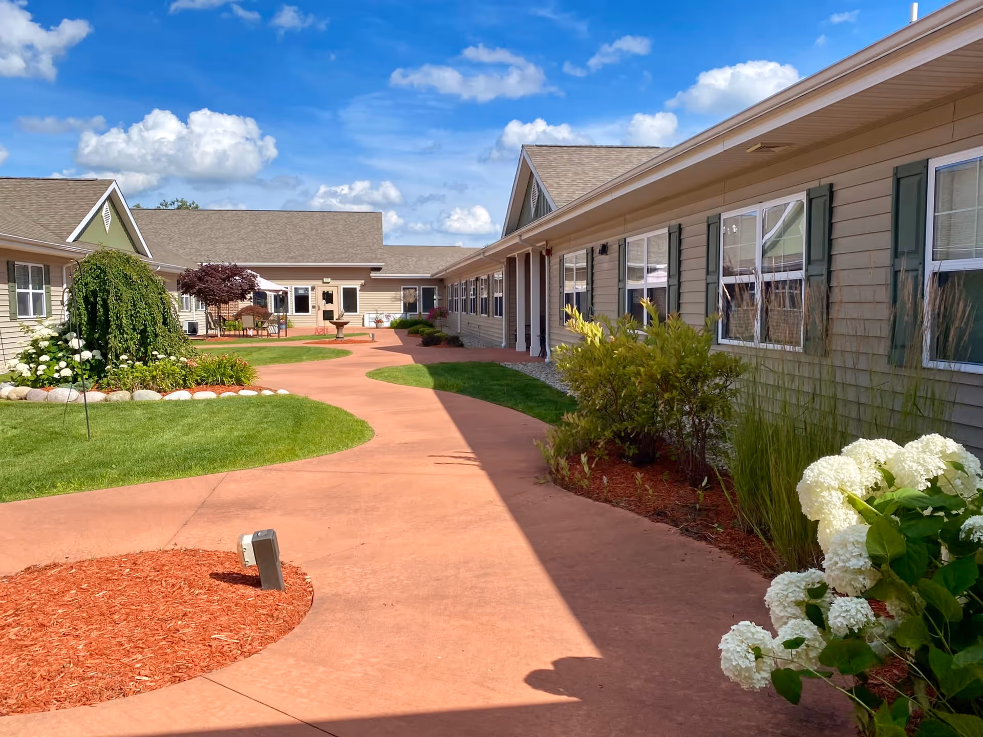 Outdoor courtyard area at Traditions of Saginaw Main Campus with a curved red walkway, green grass, various bushes and flowering plants, and beige buildings with multiple windows under a blue sky with scattered clouds.