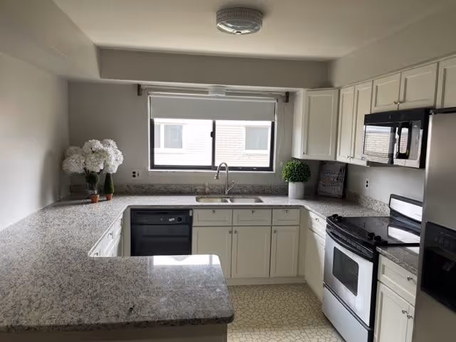 A clean and modern kitchen with white cabinets, granite countertops, a stainless steel oven and microwave, a black dishwasher, and a window above the sink letting in natural light. There are decorative plants and flowers on the countertops.