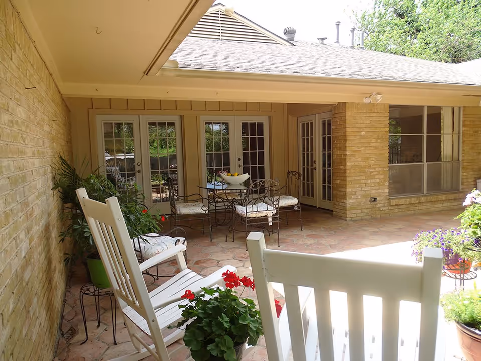 Covered outdoor patio area with white rocking chairs, potted plants with red flowers, a round metal table with four cushioned chairs, and multiple glass doors leading inside the building.