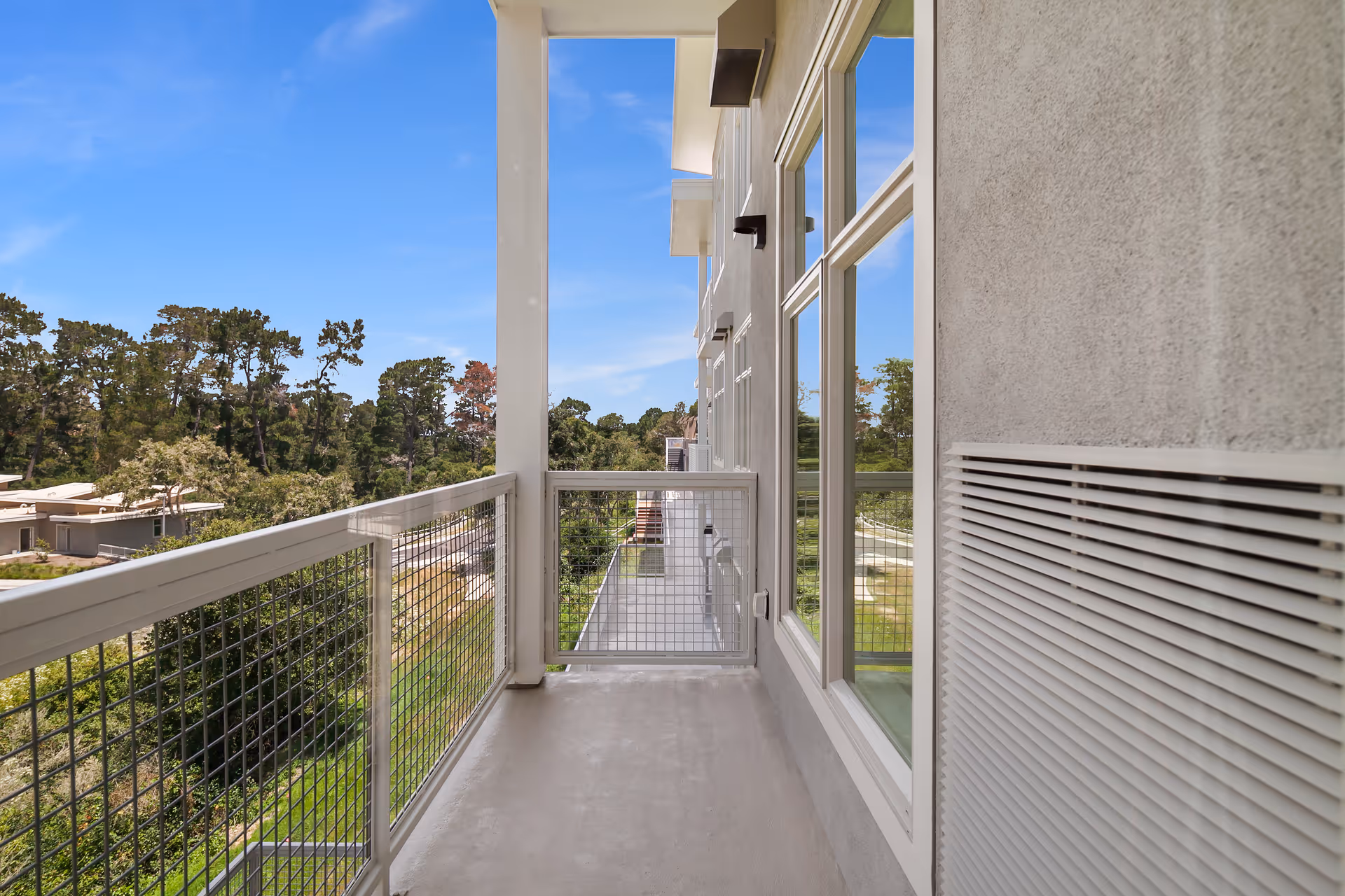 A narrow balcony with a metal railing and a small gate at the end, attached to a building with large windows. The balcony overlooks a green area with trees and other buildings under a clear blue sky.