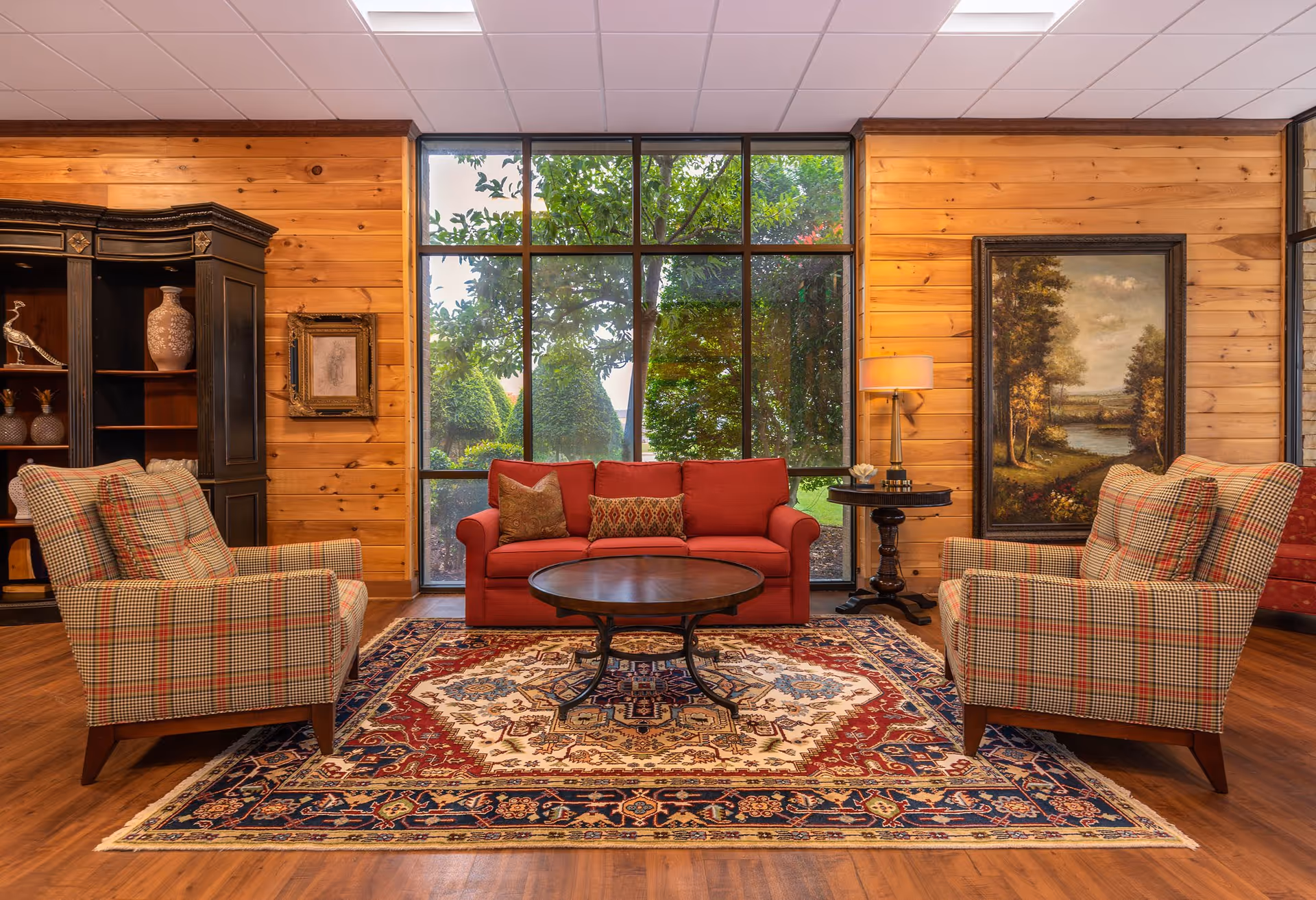 A cozy living room with wooden paneled walls, a large window showing green trees outside, a red sofa with decorative pillows, two plaid armchairs, a round wooden coffee table on a patterned rug, a black cabinet with decorative items, a side table with a lamp, and framed artwork on the walls.