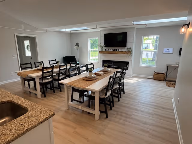 A bright and spacious dining area with two long white tables, each surrounded by black chairs. The tables are set with placemats and small decorative plants. There is a fireplace with a wooden mantel displaying the word 'OAKBROOK' beneath a mounted flat-screen TV. Two large windows allow natural light to fill the room. The floor is light wood, and a granite countertop is partially visible in the foreground.