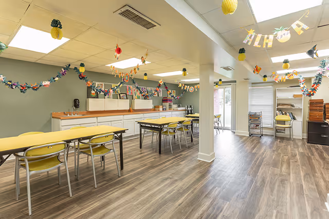 A bright and spacious room with yellow tables and chairs arranged in rows. The room has wood-patterned flooring and light green walls decorated with colorful paper garlands and hanging pineapple decorations. There are cabinets and countertops along one wall, and large windows letting in natural light.
