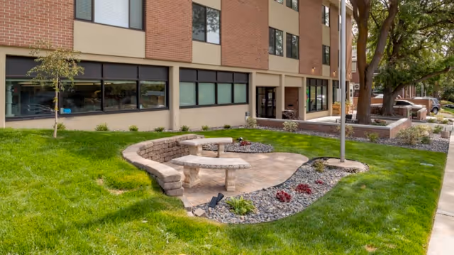 Outdoor seating area with curved stone benches and a stone table surrounded by a landscaped garden with grass, small plants, and rocks in front of a multi-story brick and beige building with large windows.