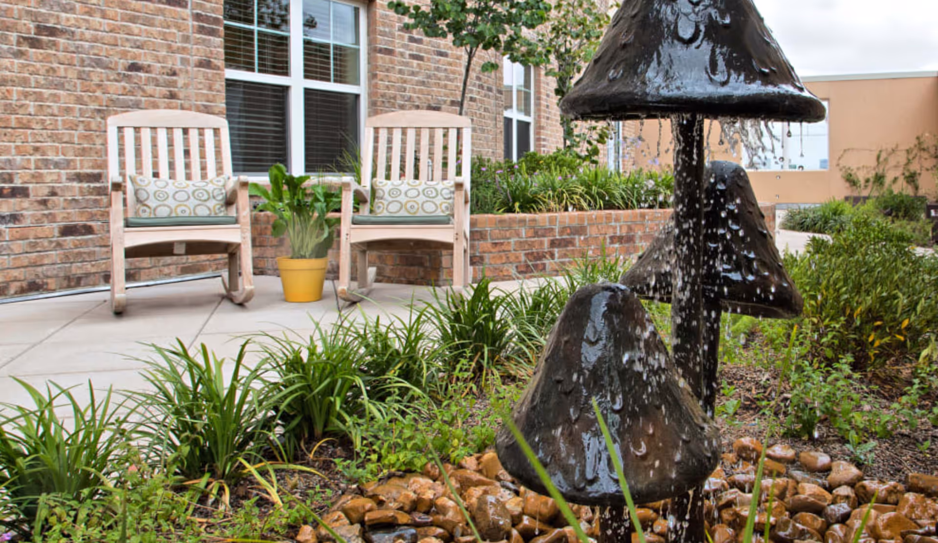 Outdoor patio area with two wooden rocking chairs, each with a patterned cushion, a yellow potted plant between them, and a decorative water fountain shaped like mushrooms with water flowing down. The background shows a brick wall with windows and some greenery.