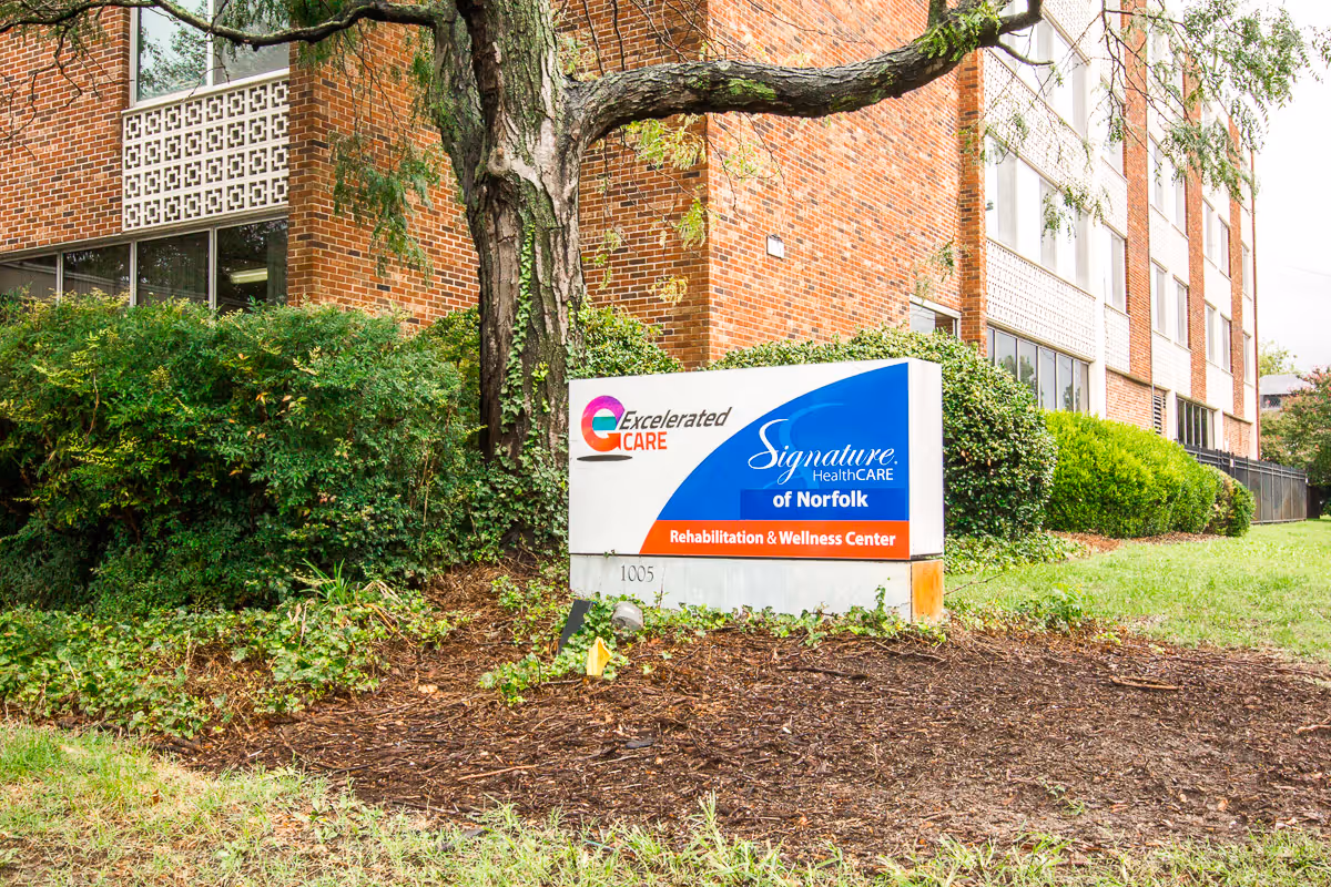Sign for Signature HealthCARE of Norfolk outside a brick rehabilitation and wellness center with trees and shrubs.
