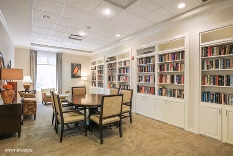 A cozy interior room featuring a round wooden table surrounded by six upholstered chairs. The room has built-in white bookshelves filled with books along one wall, beige carpeted flooring, and soft lighting from table lamps and ceiling lights. A window with curtains allows natural light to enter, and there is a piece of artwork on the wall near the window.