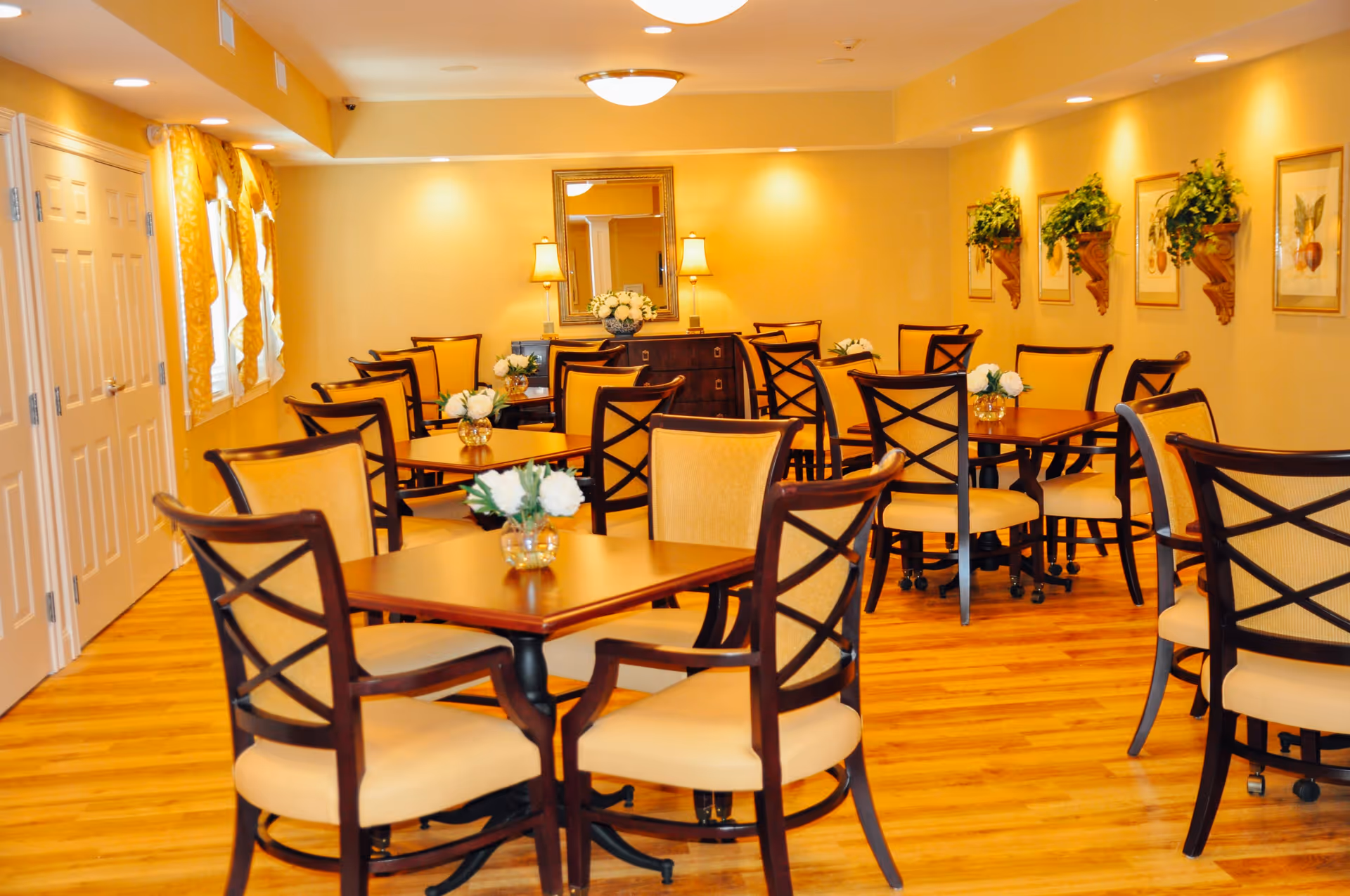 A warmly lit dining room with multiple wooden tables and cushioned chairs arranged neatly. Each table has a small vase with white flowers. The room features wooden flooring, yellow walls adorned with framed botanical prints and wall-mounted plants, and a large mirror with two lamps on a sideboard at the far end. Windows with yellow curtains are on the left side.