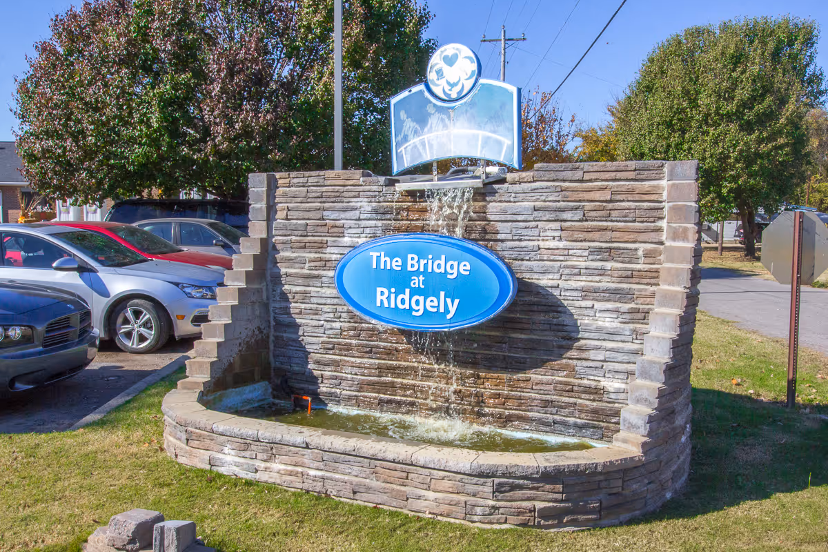 Stone fountain sign reading 'The Bridge at Ridgely' outside the facility with cars parked nearby.