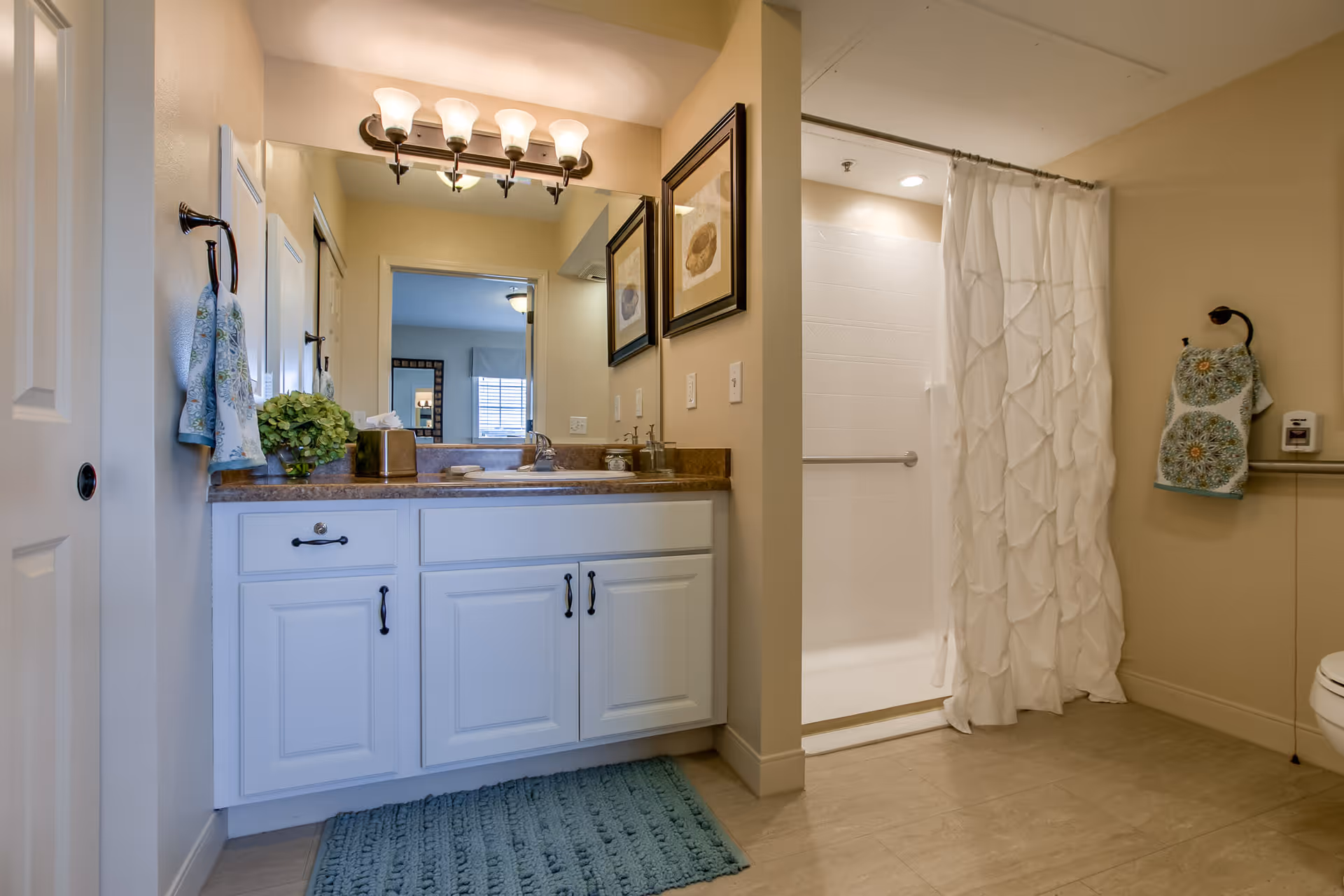 A clean and well-lit bathroom featuring a white vanity with a brown countertop, a large mirror with a light fixture above it, a shower area with a white ruffled curtain, and decorative towels hanging on the walls. The floor is tiled, and there are framed pictures on the wall next to the mirror.