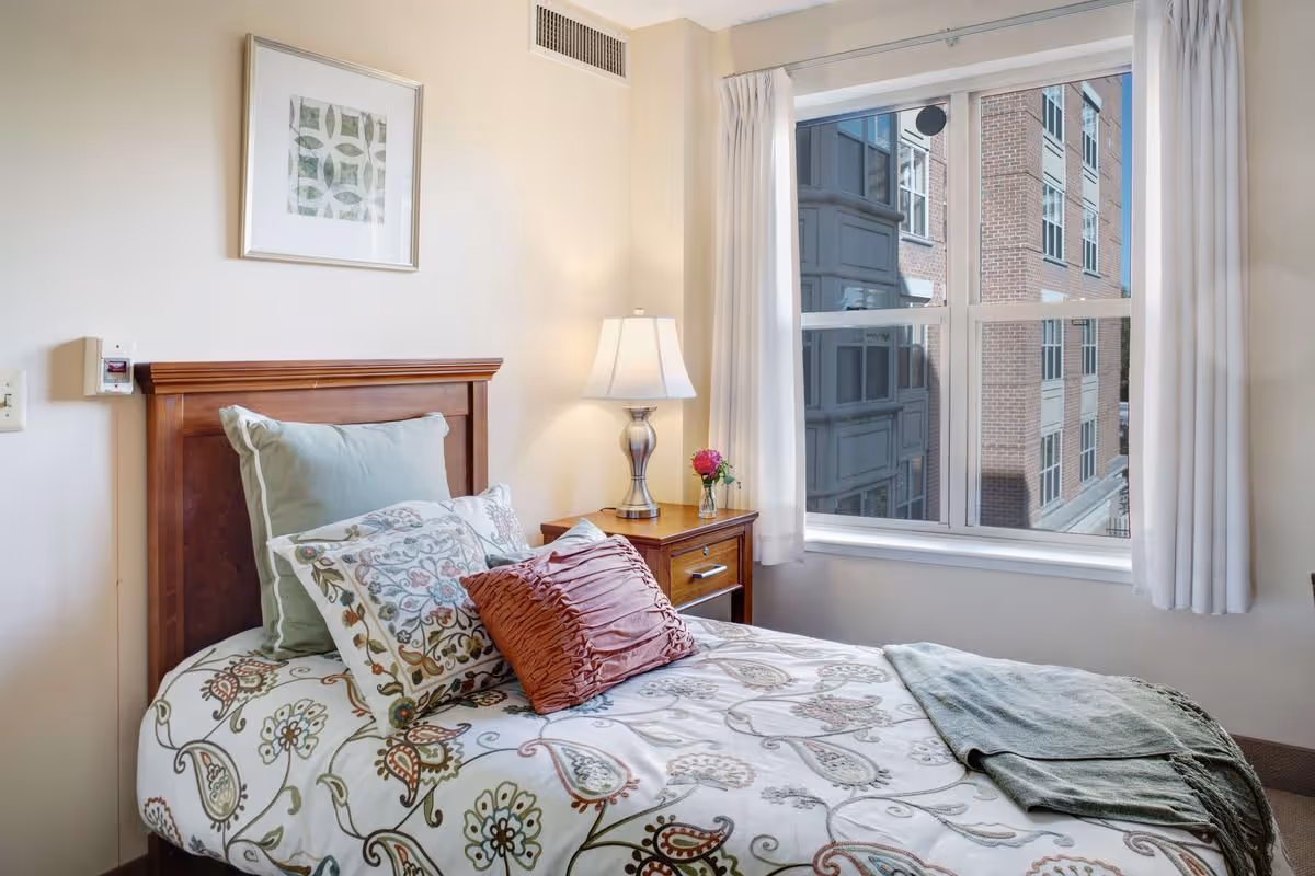 A cozy bedroom with a single bed featuring floral patterned bedding and multiple pillows, a wooden headboard, a bedside table with a lamp and a small vase with a pink flower, and a large window with white curtains showing a view of a neighboring building.