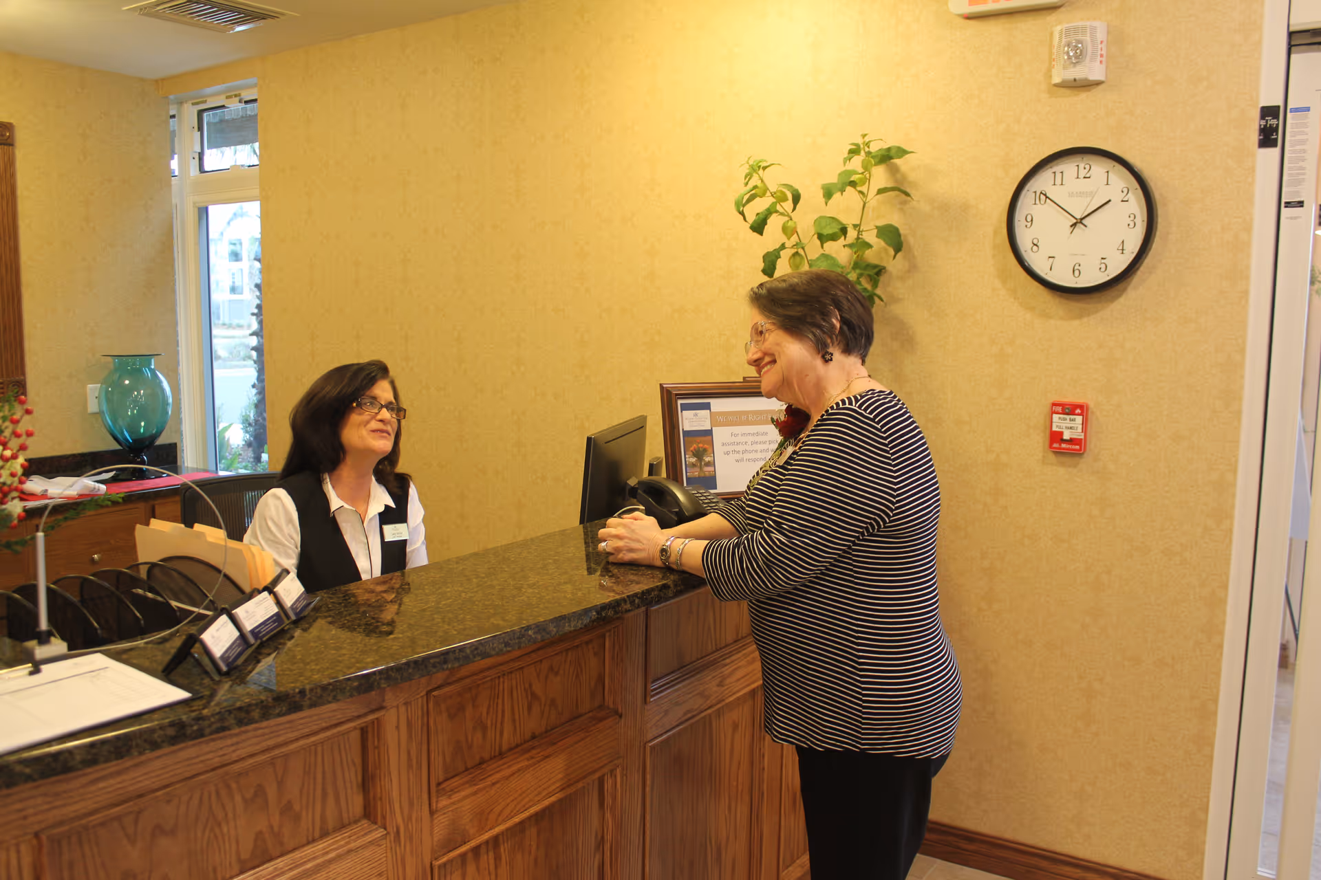 A receptionist sitting behind a wooden front desk with a granite countertop is smiling and talking to an elderly woman standing on the other side of the desk. The room has beige wallpaper, a clock on the wall showing 11:10, a fire alarm, a potted plant, and a window letting in natural light.