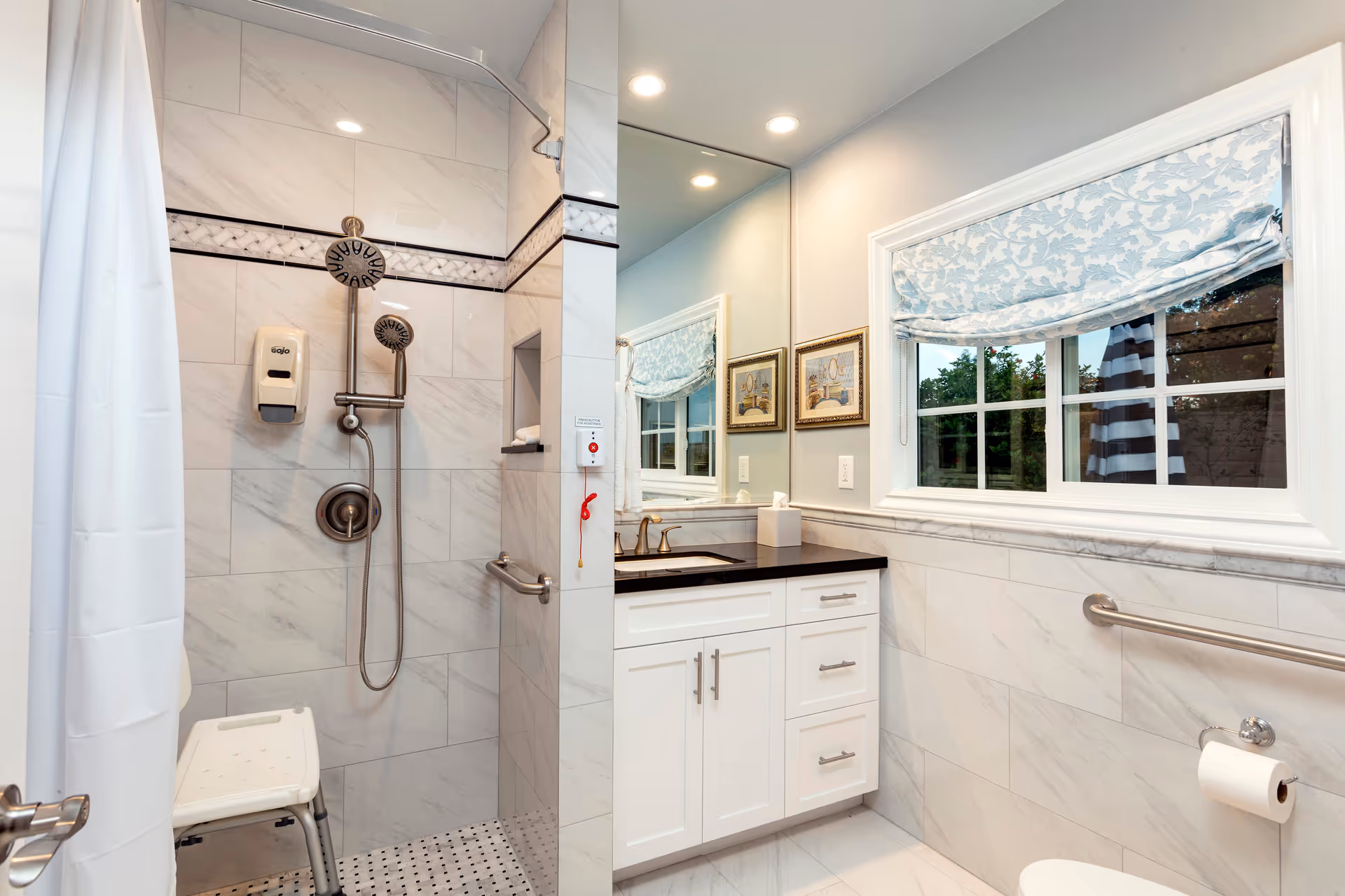 A modern bathroom with a walk-in shower featuring a handheld showerhead and a wall-mounted soap dispenser. The shower area has a white curtain and a white shower chair. The bathroom has white and gray marble tiles, a black countertop with a sink, a large mirror, two framed pictures on the wall, a window with a blue patterned Roman shade, and safety grab bars near the toilet and shower.