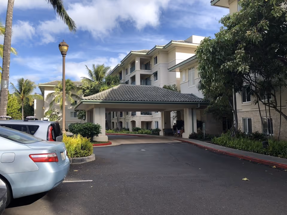 Front entrance of a multi-story senior living building with a covered porte-cochere, parked cars, palm trees, and a blue sky.
