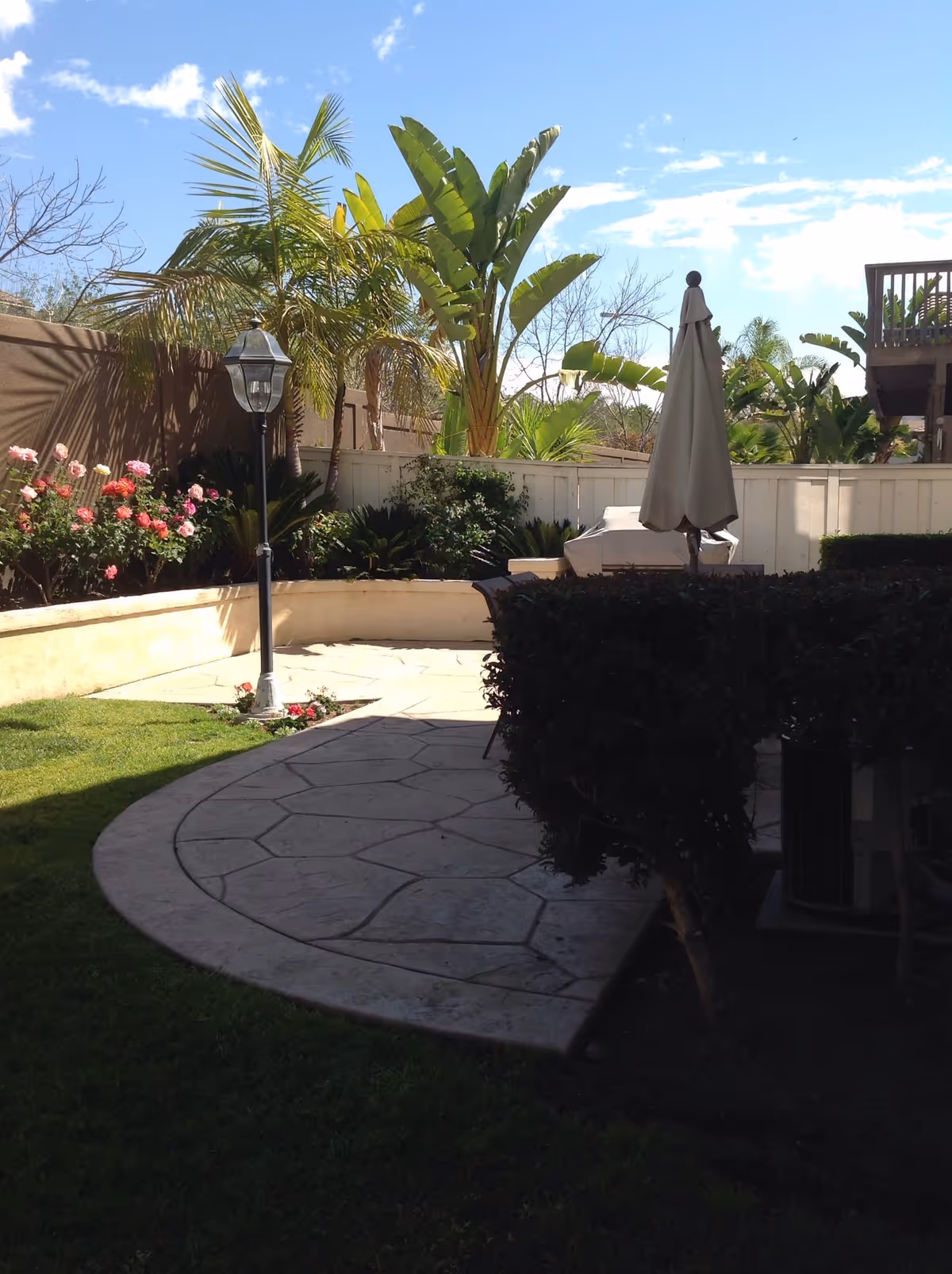 Outdoor patio area with a curved stone pathway, green grass, a lamp post, and various plants including palm trees and flowering bushes. There is a closed patio umbrella and some outdoor furniture partially visible behind a hedge. The sky is clear with some clouds.