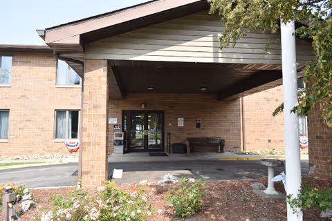 Brick senior living facility entrance with a covered porch, glass doors, and landscaped surroundings.