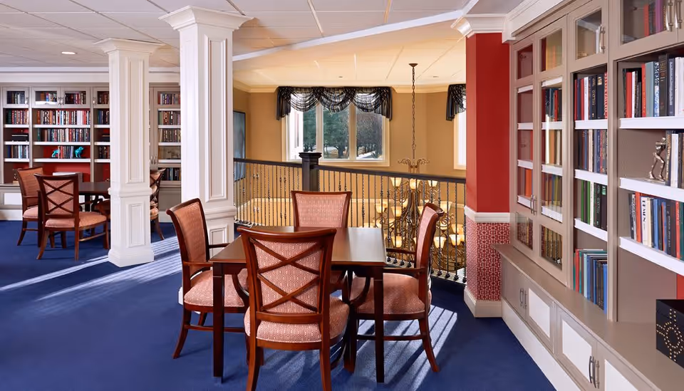 A well-lit common area with blue carpet, wooden tables and chairs with red upholstery, and built-in bookshelves filled with books. The space features white columns, large windows with decorative curtains, and a chandelier hanging over a stairwell.