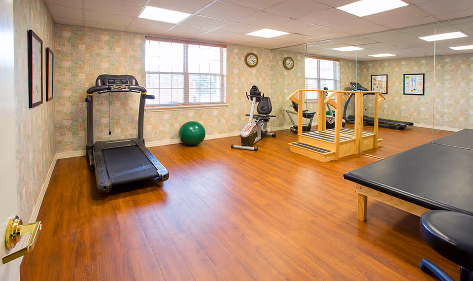 A fitness room with wooden flooring and patterned wallpaper. The room contains a treadmill, an exercise bike, a green exercise ball, a wooden balance ramp, and a padded therapy table. Two windows with blinds allow natural light into the room, and two clocks are mounted on the wall between the windows. Framed pictures hang on the walls.